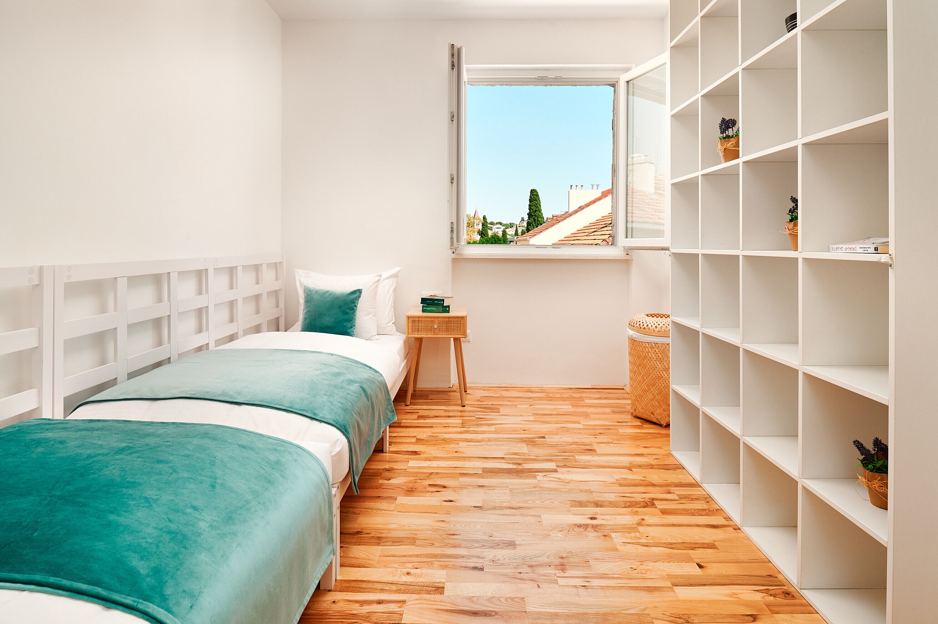 A small, bright room with two beds, a window, and a white shelving unit.