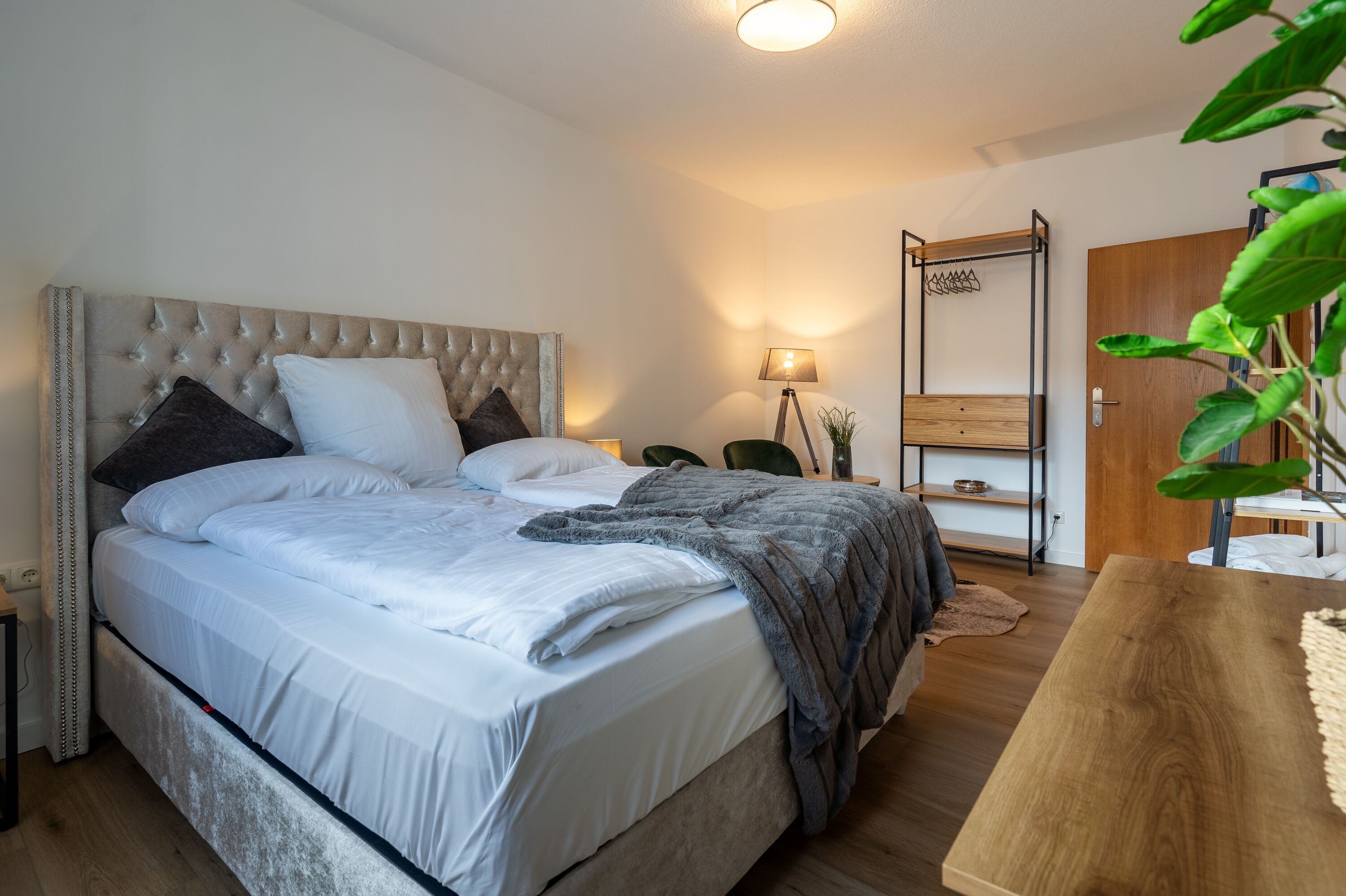Ein Schlafzimmer mit einem gesteppten Kopfteil, einem Bett mit weißen Bettbezügen, einer grauen Tagesdecke, einem Nachttisch, einer Lampe, einem Holzregal und einer Holztür.