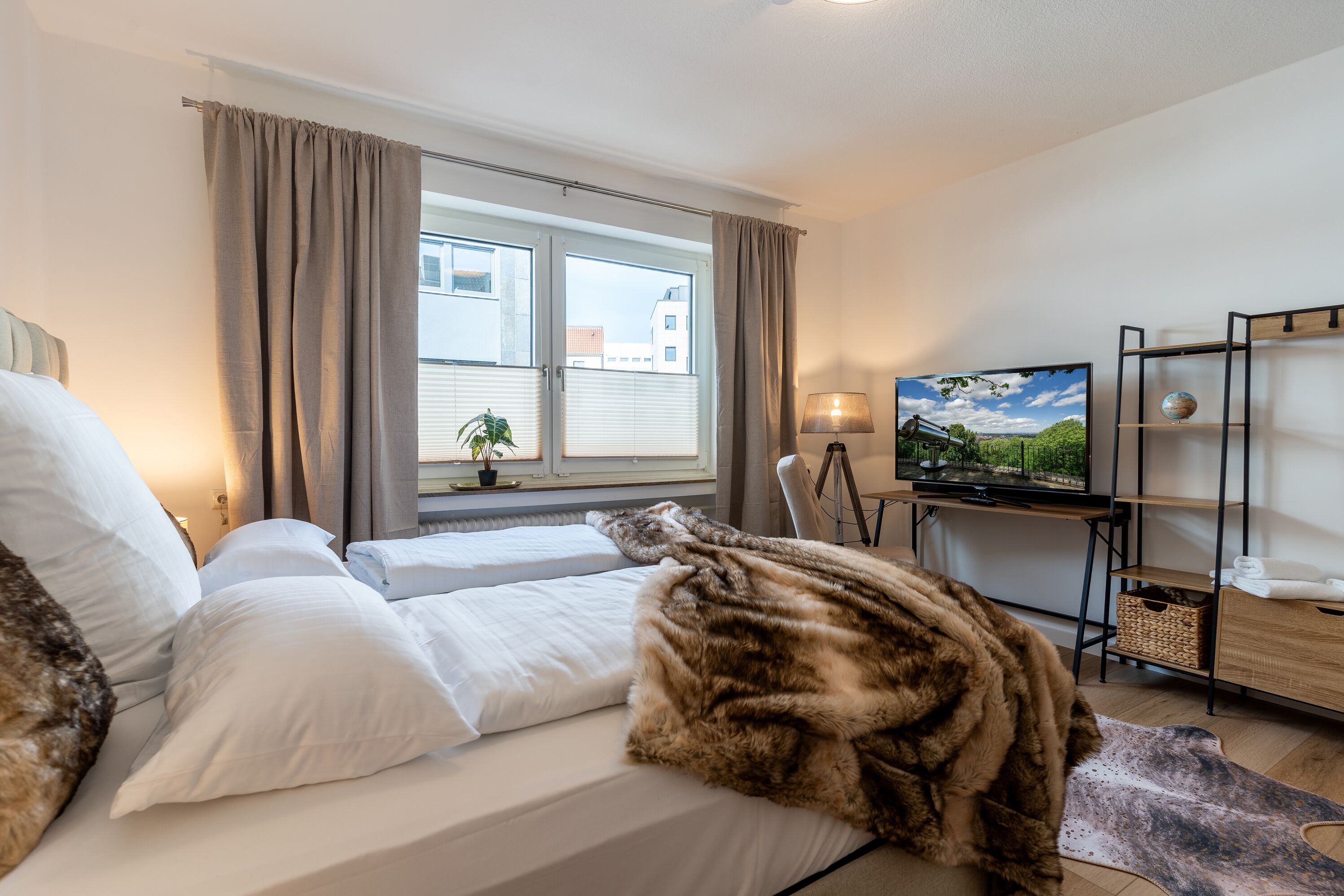 A hotel room with a bed, a TV, a desk with a lamp, and a window with curtains.