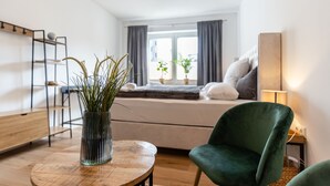 Un dormitorio moderno con cama, una silla verde, una mesa redonda de madera con un florero, y una ventana con cortinas.