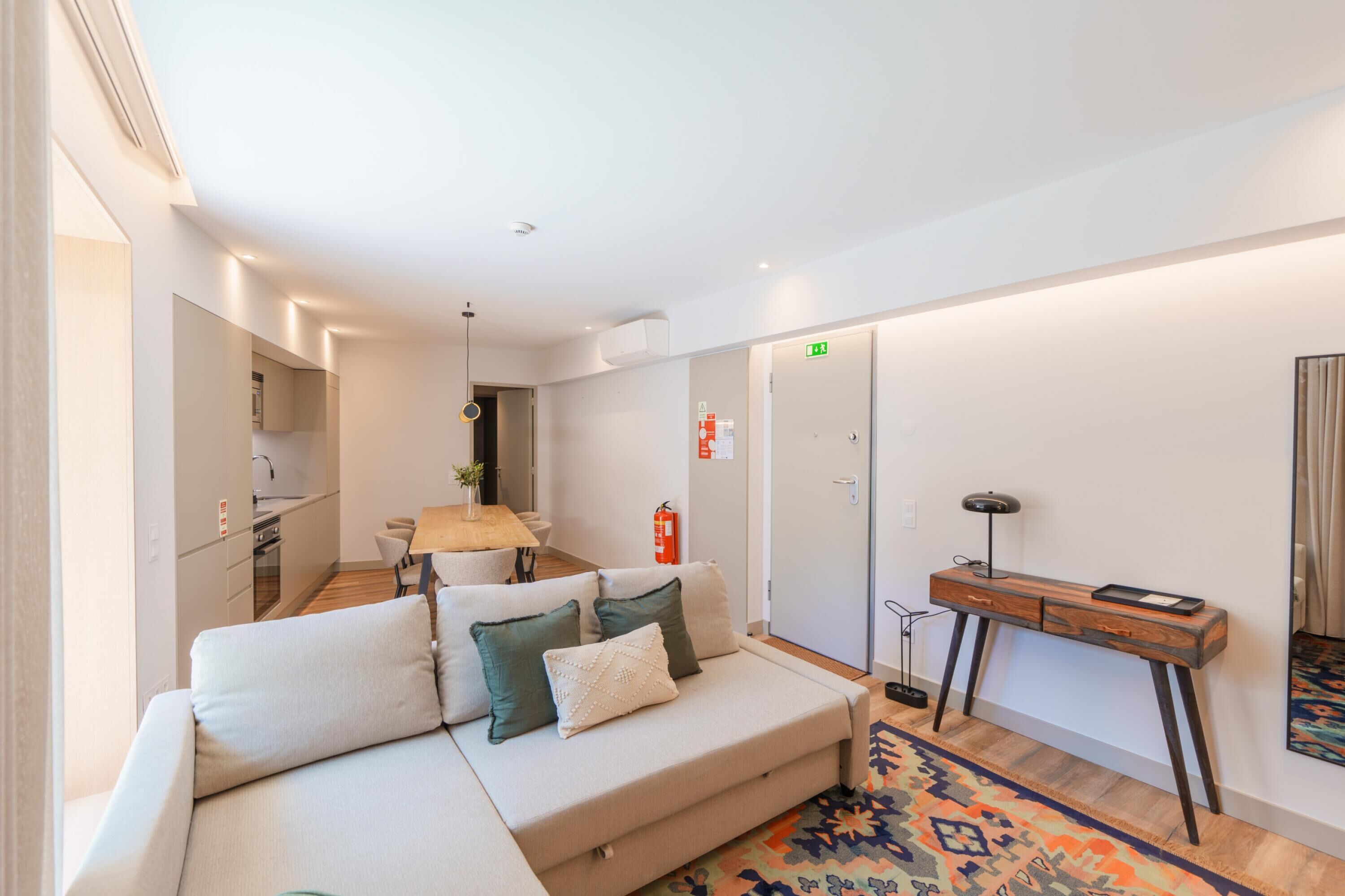 A modern hotel room with a sofa, a wooden desk, a patterned rug, and a kitchenette in the background.