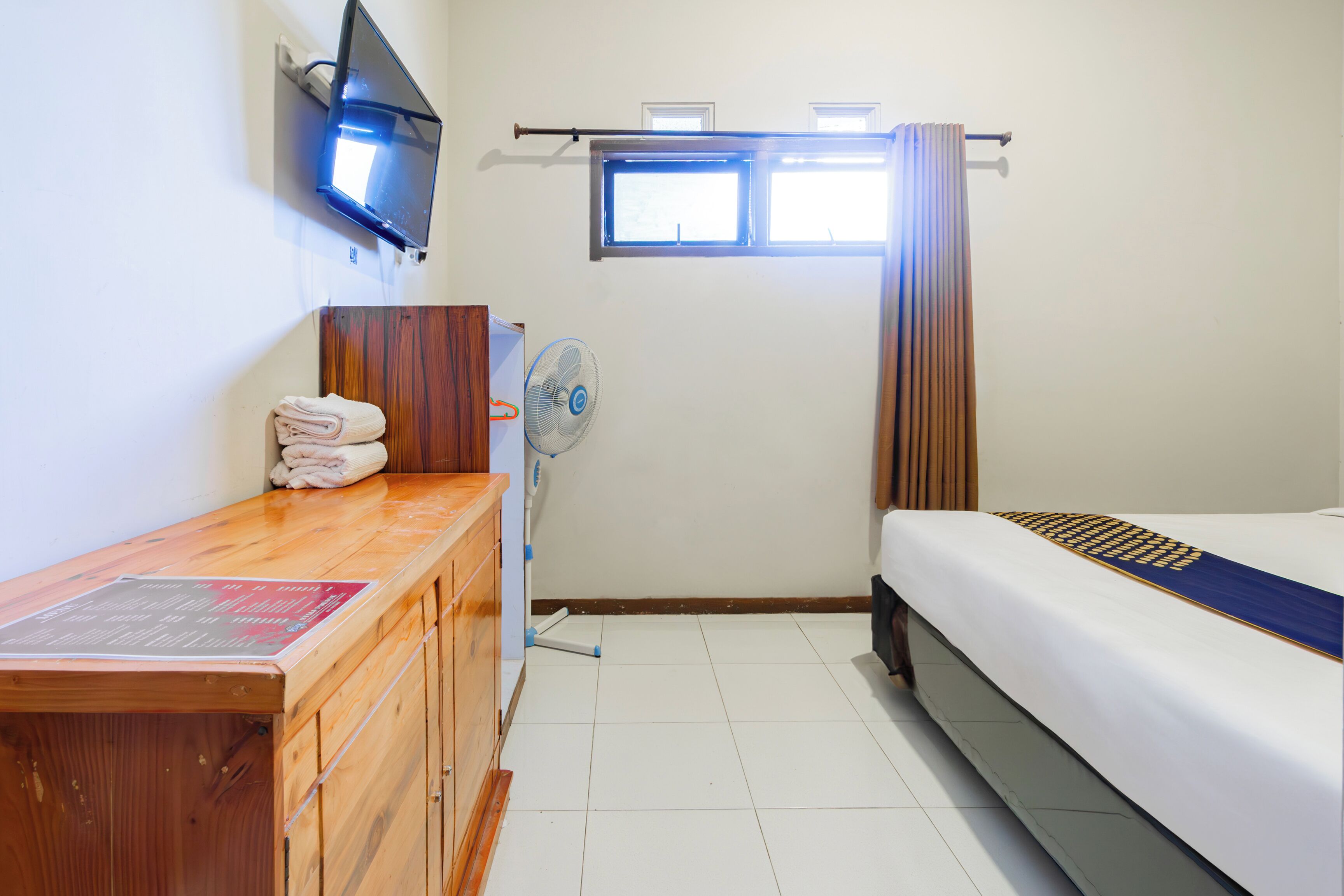 A hotel room with a wooden desk, a bed, a TV mounted on the wall, and a window with curtains.