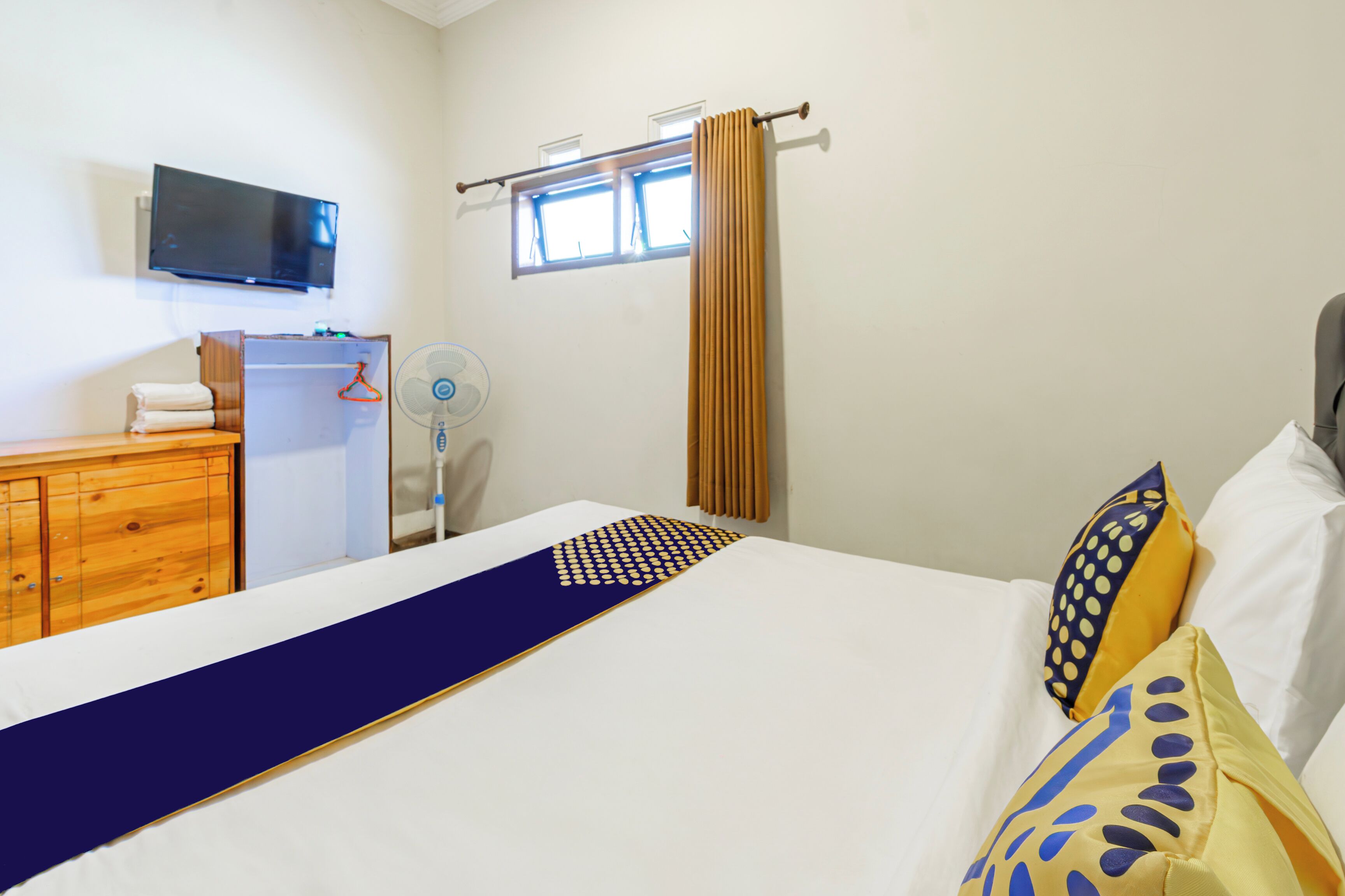 A hotel room with a bed, a TV mounted on the wall, a window with curtains, a wooden dresser, and a fan.