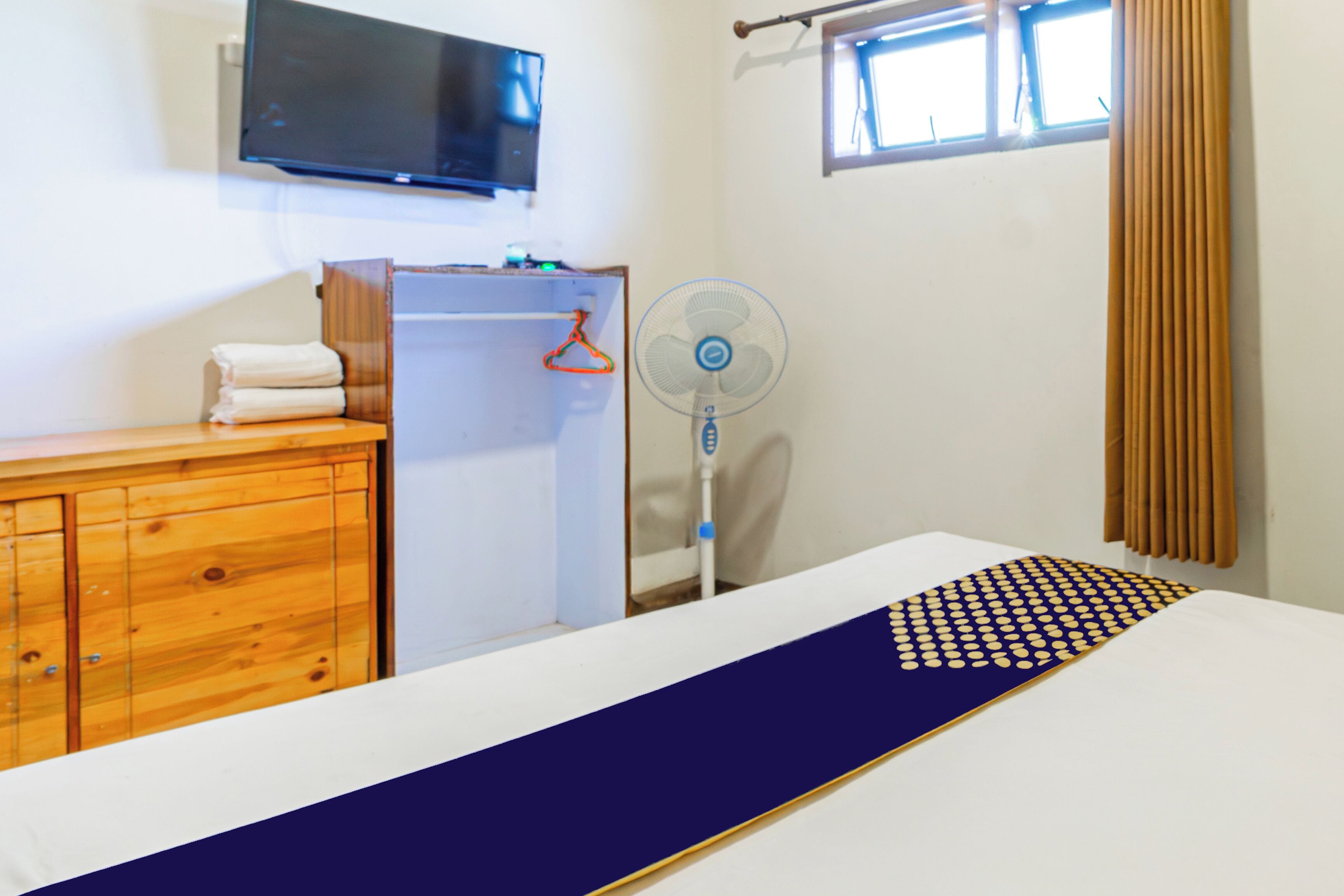 Une chambre d’hôtel avec un lit, une commode en bois, un téléviseur fixé au mur, une fenêtre avec des rideaux et un ventilateur sur pied.
