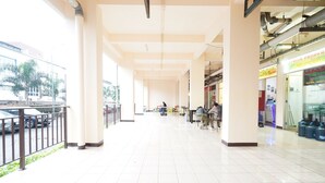 A spacious corridor with tiled floors, large pillars, and a railing on one side. There are several shops on the right, including one with a sign that reads "JANGIN PROPERTIES". - Hotel O Triple N Homestay (Sidoarjo)