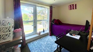 Une chambre avec un lit, un bureau avec un ordinateur portable, une fenêtre avec des rideaux et un petit chien près de la fenêtre.