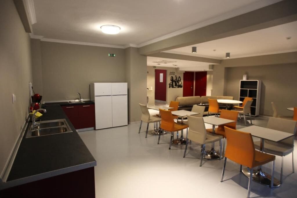Un espace repas moderne avec des tables et des chaises, un coin cuisine équipé d’un réfrigérateur, et une porte rouge en arrière-plan.