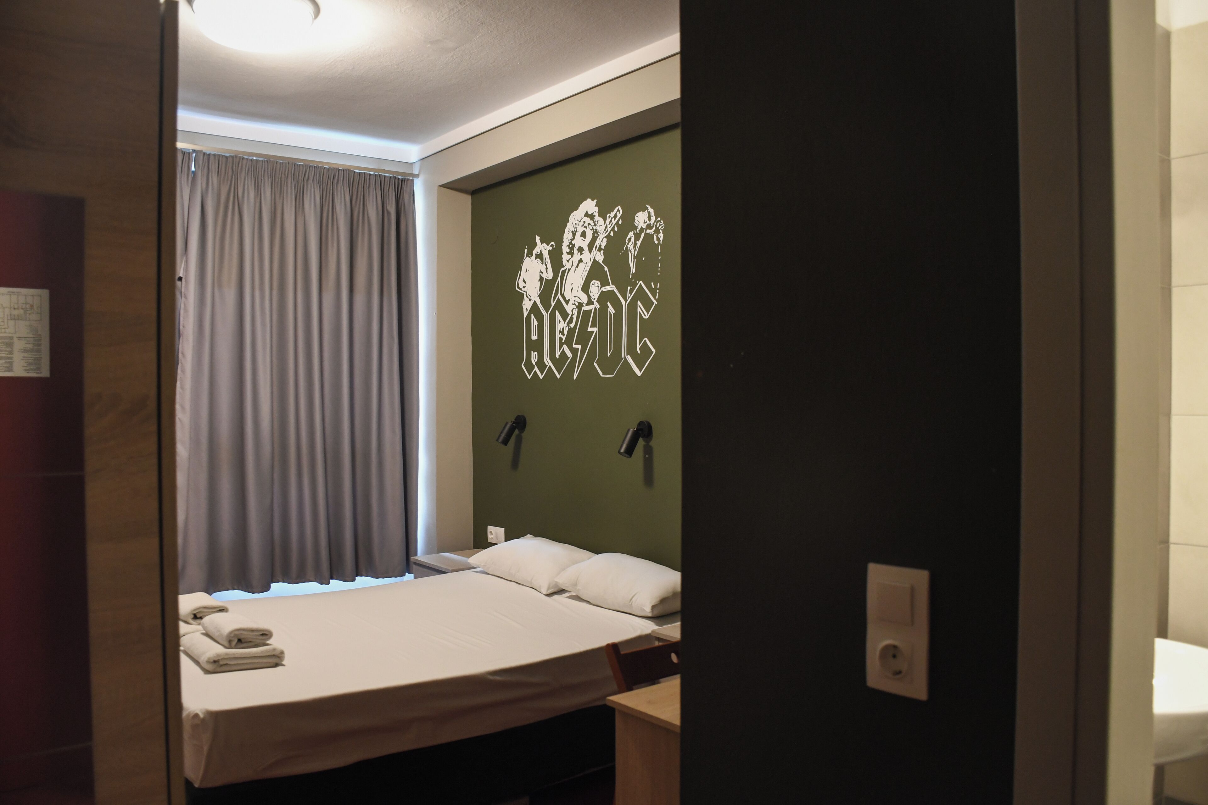Une chambre d’hôtel avec un lit, une tête de lit ornée du logo d’un groupe de musique, une fenêtre avec des rideaux et un luminaire fixé au mur.
