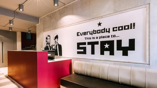 A hotel reception area with a modern design, including a red reception desk, a large wall poster with motivational text, and a seating area with a white sofa.