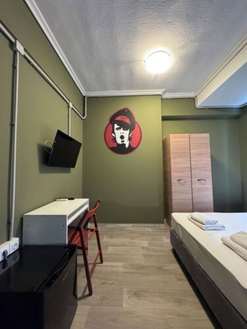 A small, compact room with a bed, a desk, a chair, a wardrobe, and a wall-mounted TV.