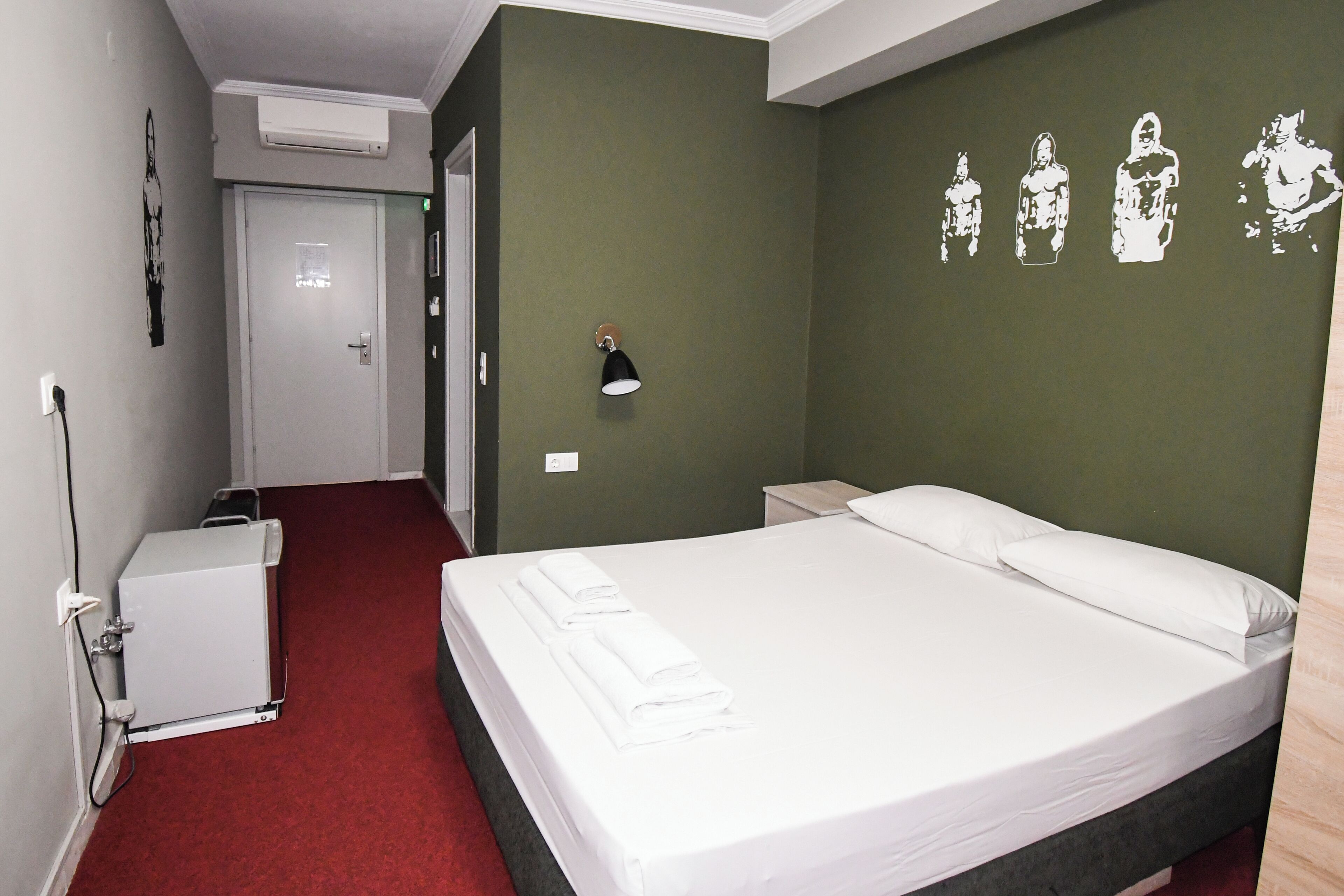 Una habitació d'hotel amb un llit, una tauleta de nit, una làmpada penjada a la paret i un terra cobert de catifes vermelles.