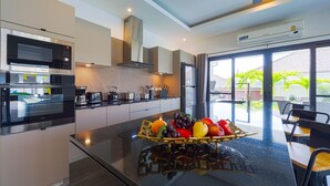 A modern kitchen with built-in appliances, a central island with a fruit bowl, and large windows offering a view of the outdoors. - Modern 3 Bedroom Private Pool Villa -ML7 (Hua Hin)