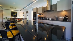 A modern kitchen with a central island, built-in oven, microwave, and stainless steel appliances. There are bar stools and a dining table in the adjacent area. - Modern 3 Bedroom Private Pool Villa -ML7 (Hua Hin)