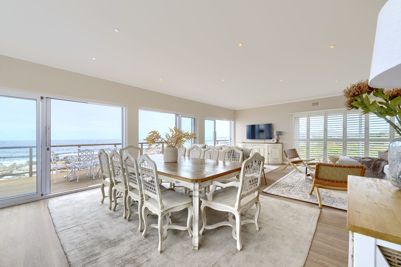 Ett rymligt matområde med ett stort träbord och vita stolar, med utsikt över en strand.
