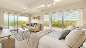 Un dormitorio con una cama grande, un sofá, una mesita de centro y un escritorio. Cuenta con ventanas amplias con persianas y vistas a zonas verdes.