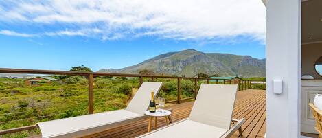 A wooden deck with two lounge chairs and a table, overlooking a mountainous landscape.