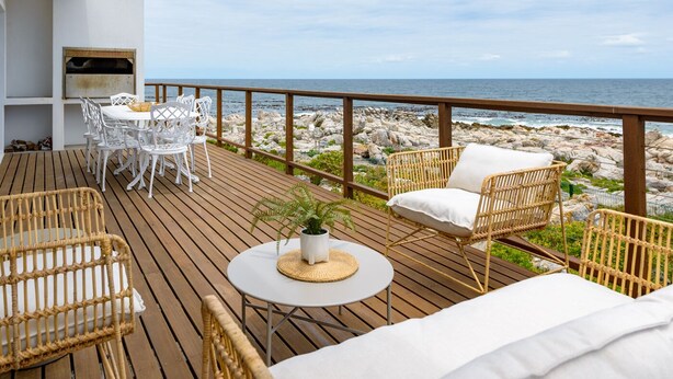 Una terraza de madera con mobiliario exterior, incluyendo sillas de mimbre y una mesa, con vistas a una costa rocosa.
