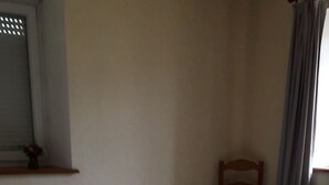 A bedroom with a bed, a chair, a small table with a lamp, and a window with curtains.