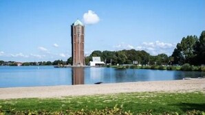 Una torre junto a un lago tranquilo, con un primer plano de hierba y árboles al fondo.