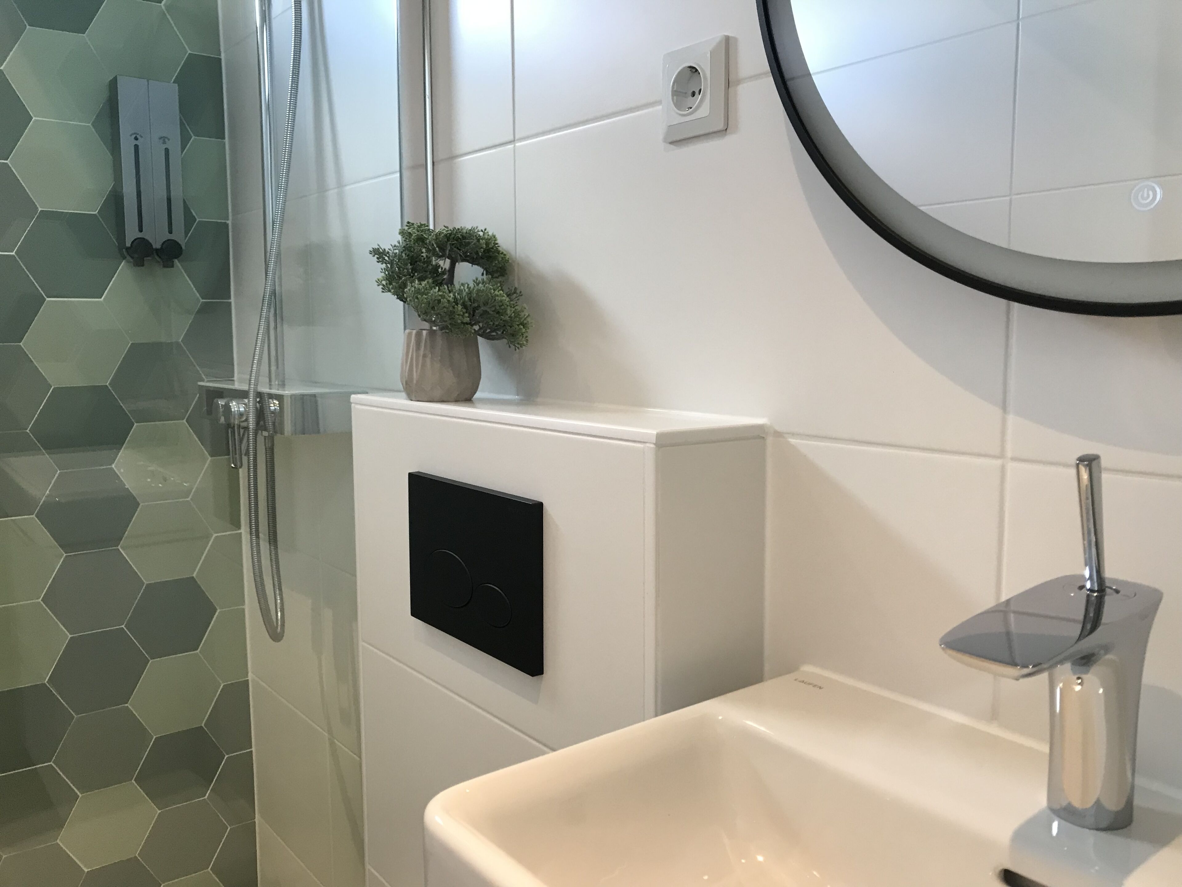 Un baño con cabina de ducha de vidrio, un lavabo blanco con grifo moderno, una planta en maceta en la pared y un espejo redondo.