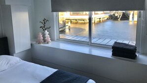 Habitación de hotel con cama, ventana que ofrece vista a un muelle y barcos, un pequeño florero con ramas y una toalla doblada en el alféizar.