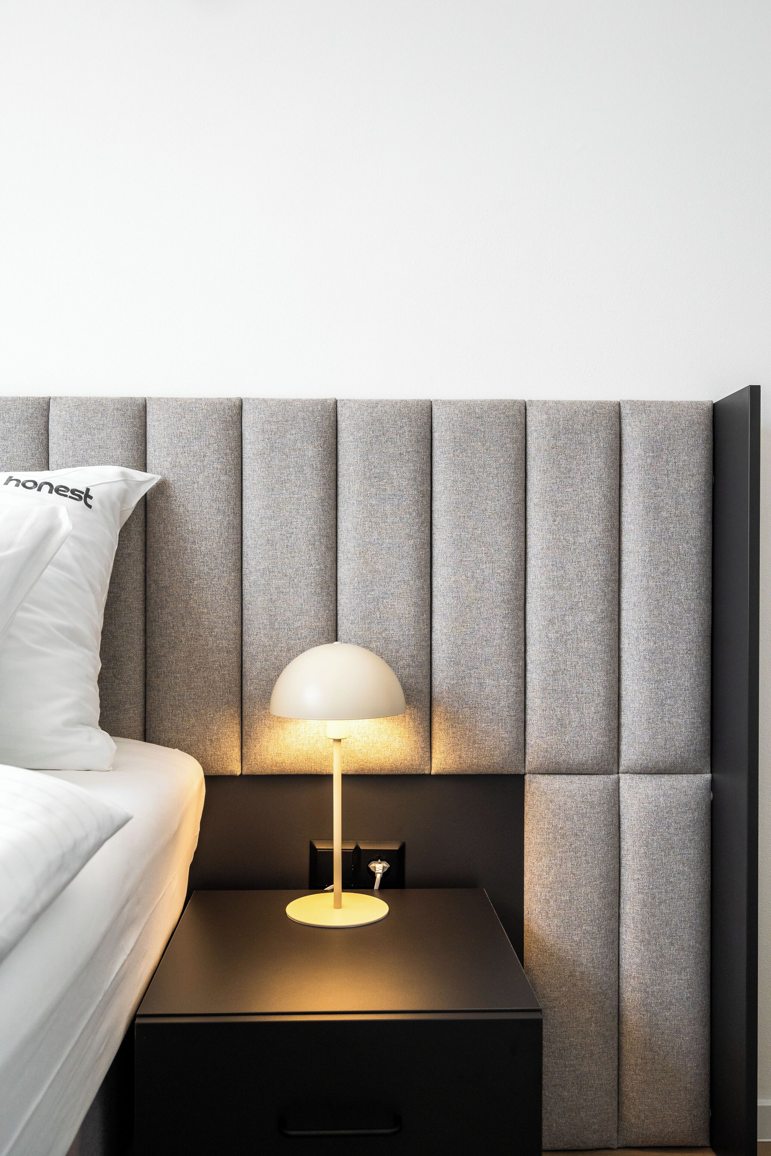 Un dormitorio moderno con un cabecero tapizado en gris, una mesita de noche con una lámpara y una almohada blanca con la palabra 'honest' escrita.
