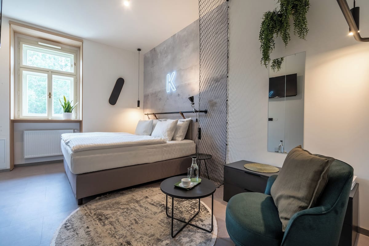 A modern bedroom with a bed, bedside table, and a chair. There is a window with a plant, a wall-mounted TV, and a neon 'K' sign.