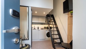 Een kleine keuken met witte kasten, een wasmachine en een koffiezetapparaat. Een zwarte trap staat tegen de muur, en boven het aanrecht is een televisie bevestigd.