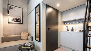 Een moderne kamer met een bank, een klein tafeltje met een lamp, een lijst met kunstwerk aan de muur, een donkere deur, en een keukenhoek met kastjes en een spoelbak.