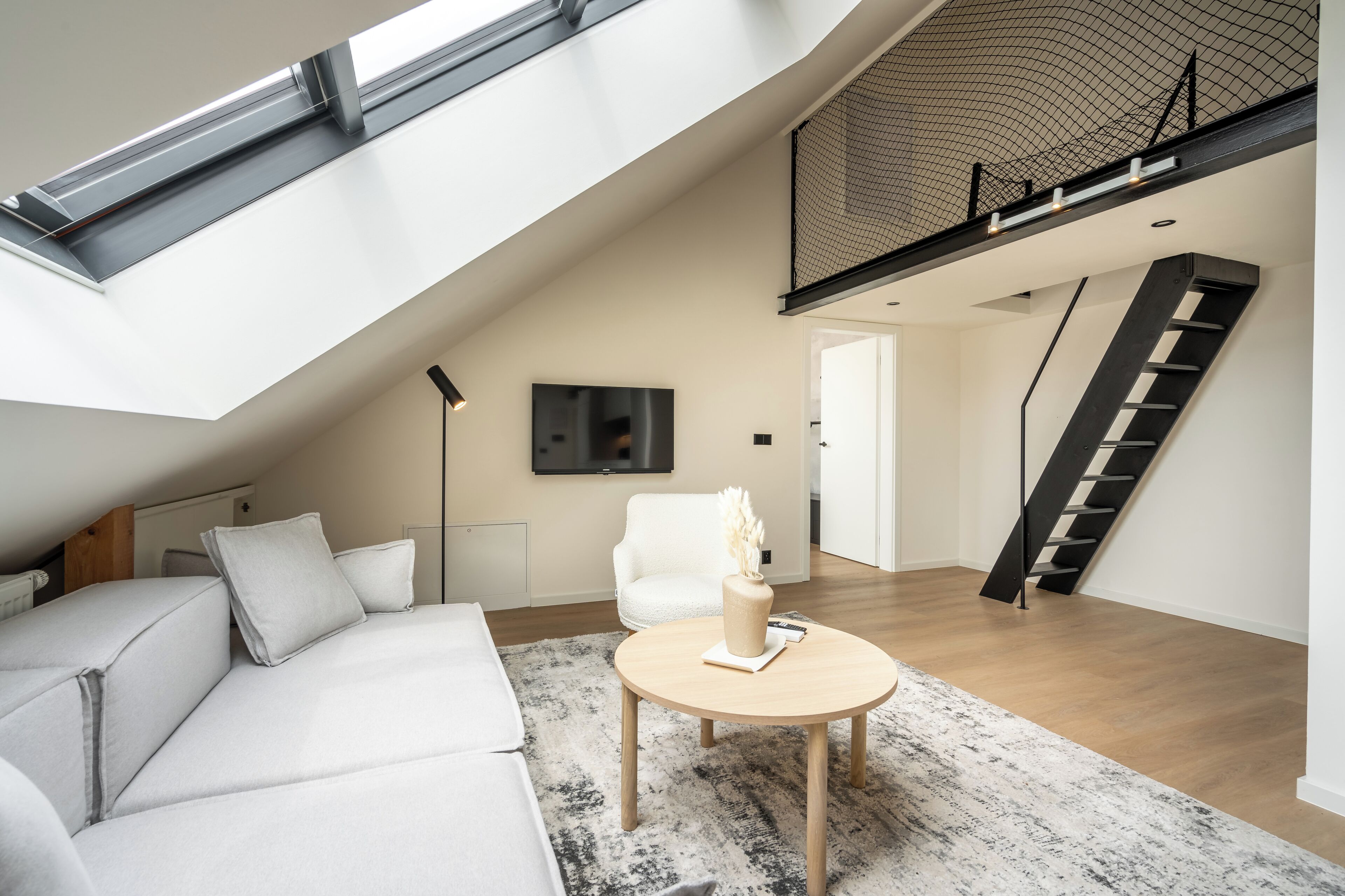 A modern loft apartment with a high ceiling, a skylight, a white sofa, a round coffee table, and a staircase leading to an upper level.