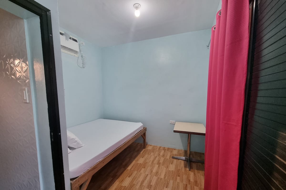 Una habitación peque ña con cama individual, piso de madera, una mesita y una cortina rosa.