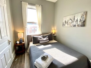 A neatly arranged bedroom with a bed, bedside lamps, a window with curtains, and a cityscape painting on the wall.