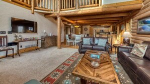 A spacious log cabin interior with a high ceiling, wooden beams, a cozy living area with a sofa and coffee table, and a kitchen visible in the background. - ********River Cabin Minutes To Town***** (Hamilton)