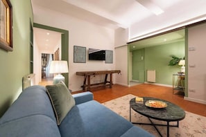 A living room with a blue sofa, a coffee table, a TV mounted on the wall, and a dining area visible through an open door.