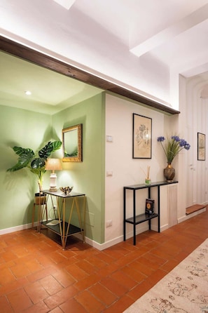 A hallway with a green wall, a wooden ceiling beam, a table with a lamp, a potted plant, and a console table with a vase.