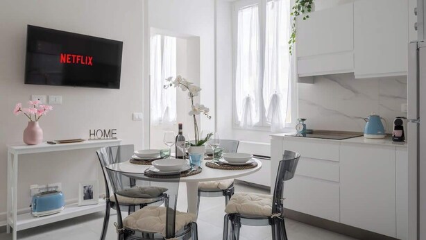 Une cuisine moderne avec des meubles blancs, une table à manger entourée de chaises, un téléviseur affichant le logo « NETFLIX », et un vase rempli de fleurs posé sur une étagère.