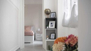 Un couloir mène à une chambre à coucher qui contient un lit, une table de chevet, une lampe, une étagère avec des photos encadrées, un globe terrestre et un vase avec des fleurs.