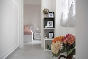 A hallway leading to a bedroom with a bed, a nightstand, a lamp, a shelf with framed pictures, a globe, and a vase with flowers.