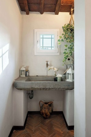 A narrow bathroom with a stone sink, a window, a hanging plant, and a rustic vase.