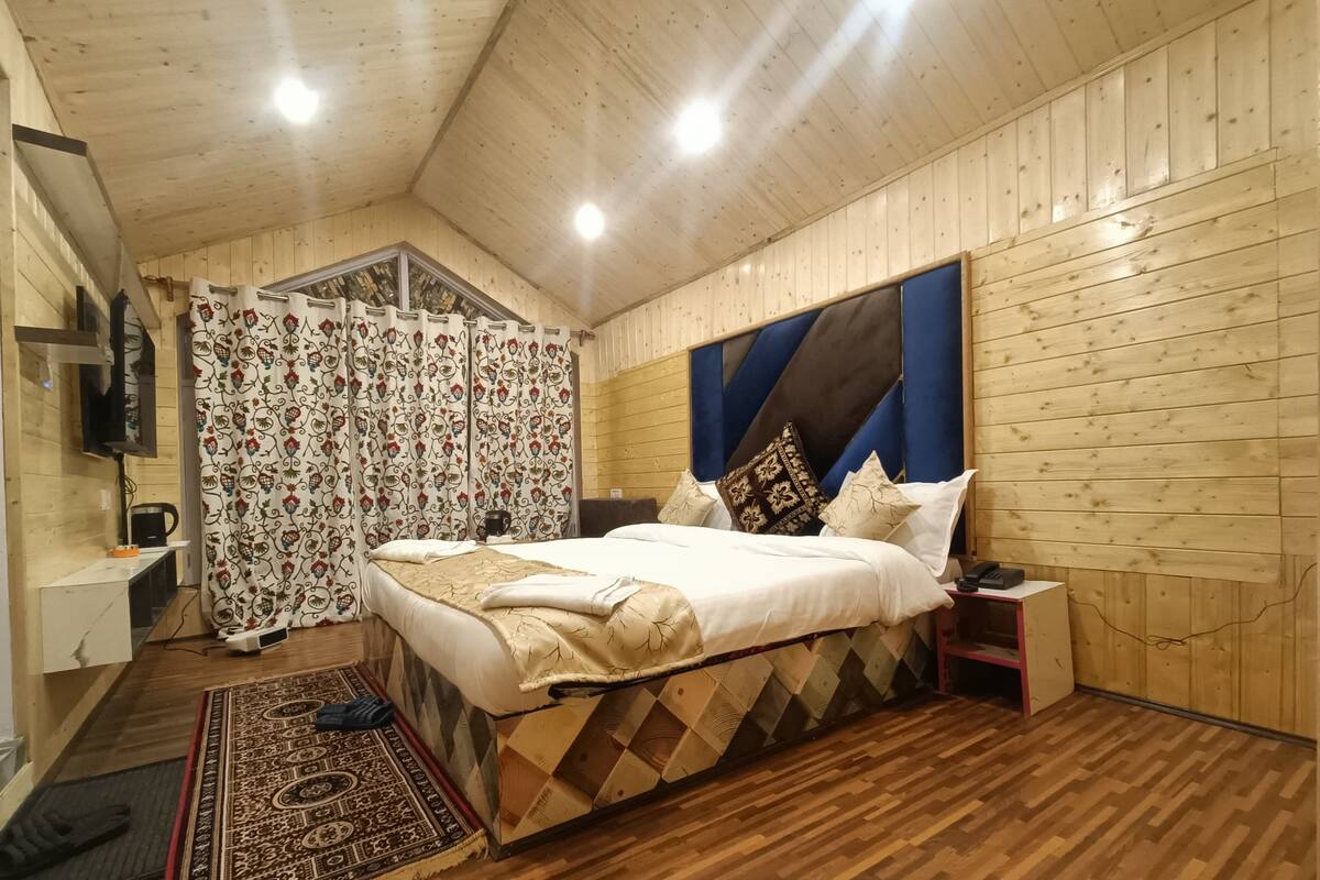 A cozy bedroom with a wooden ceiling, a large bed with patterned bedding, a TV mounted on the wall, and a patterned rug on the floor.