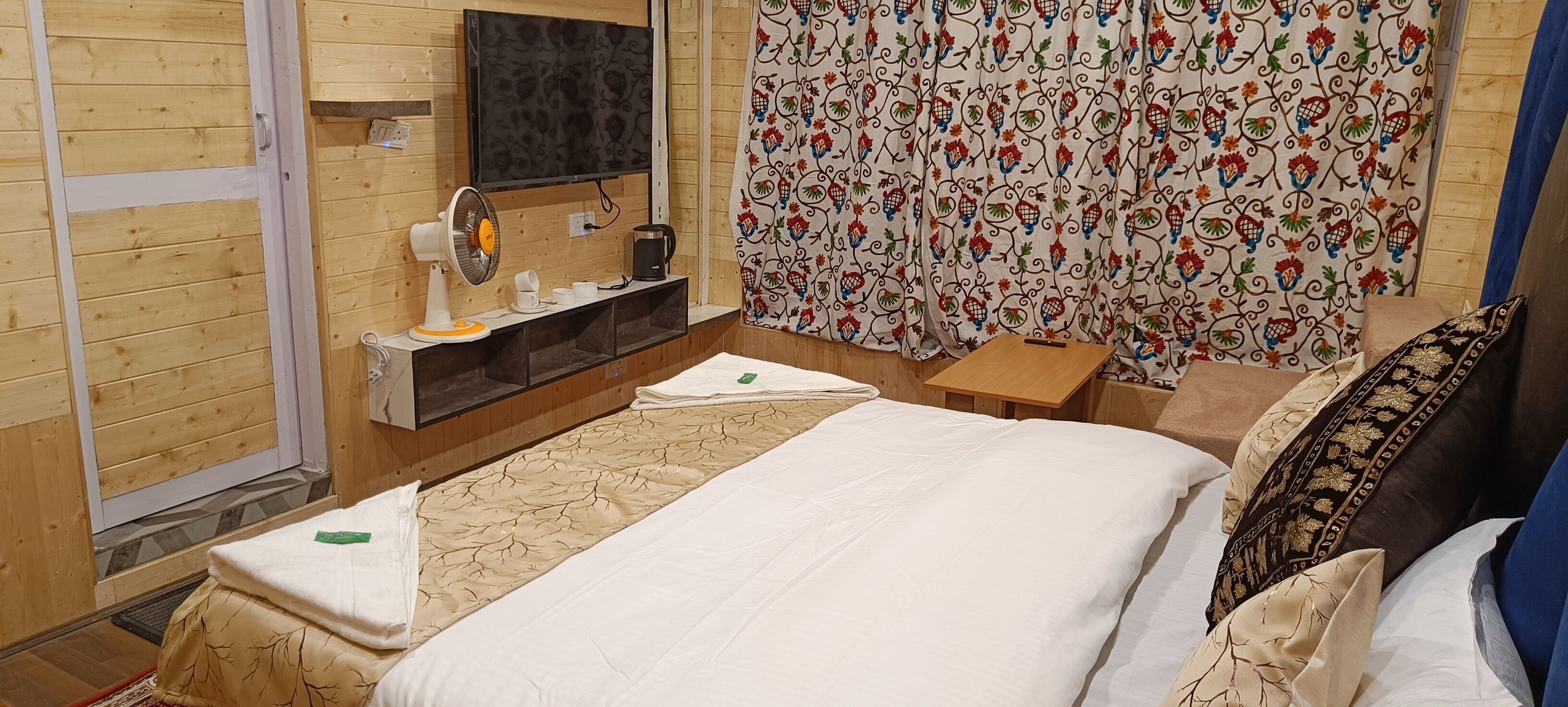 Habitación de hotel con cama, mesita de noche, televisor, ventilador y ventana con cortinas.