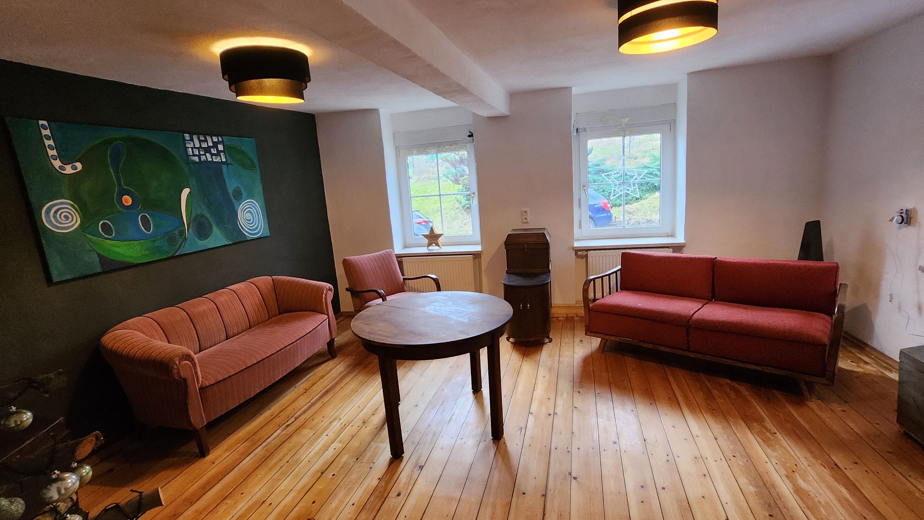 Un salon avec un plancher en bois, un canapé rouge, une table ronde et un tableau accroché au mur.