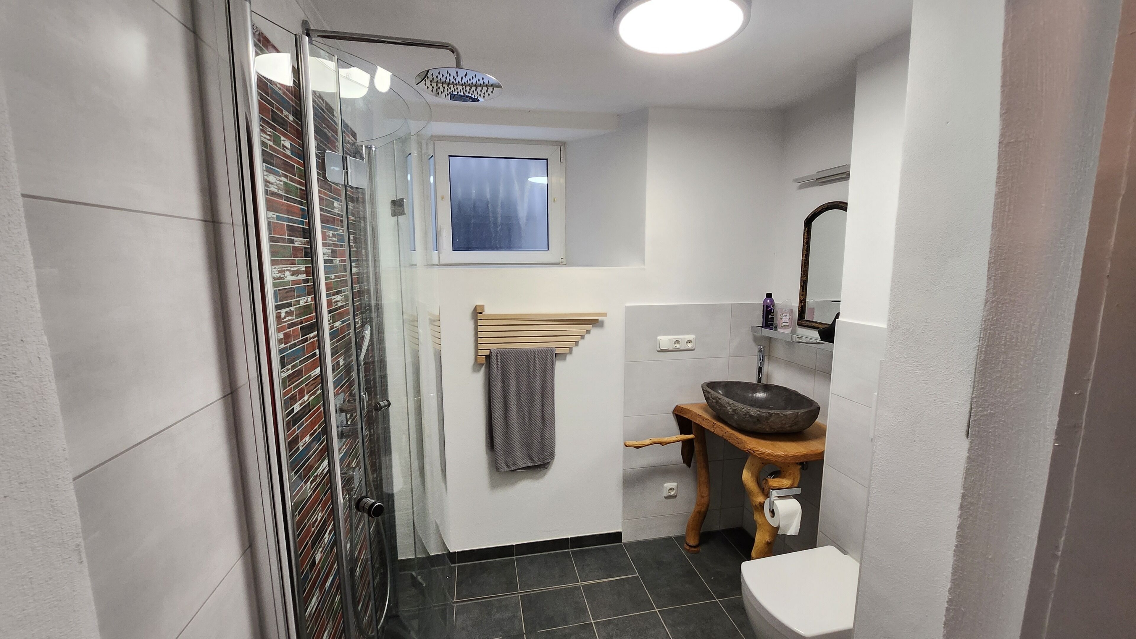 Une petite salle de bain dotée d’une douche à parois de verre, d’un porte-serviettes en bois, d’un lavabo avec un receveur en pierre et de toilettes.