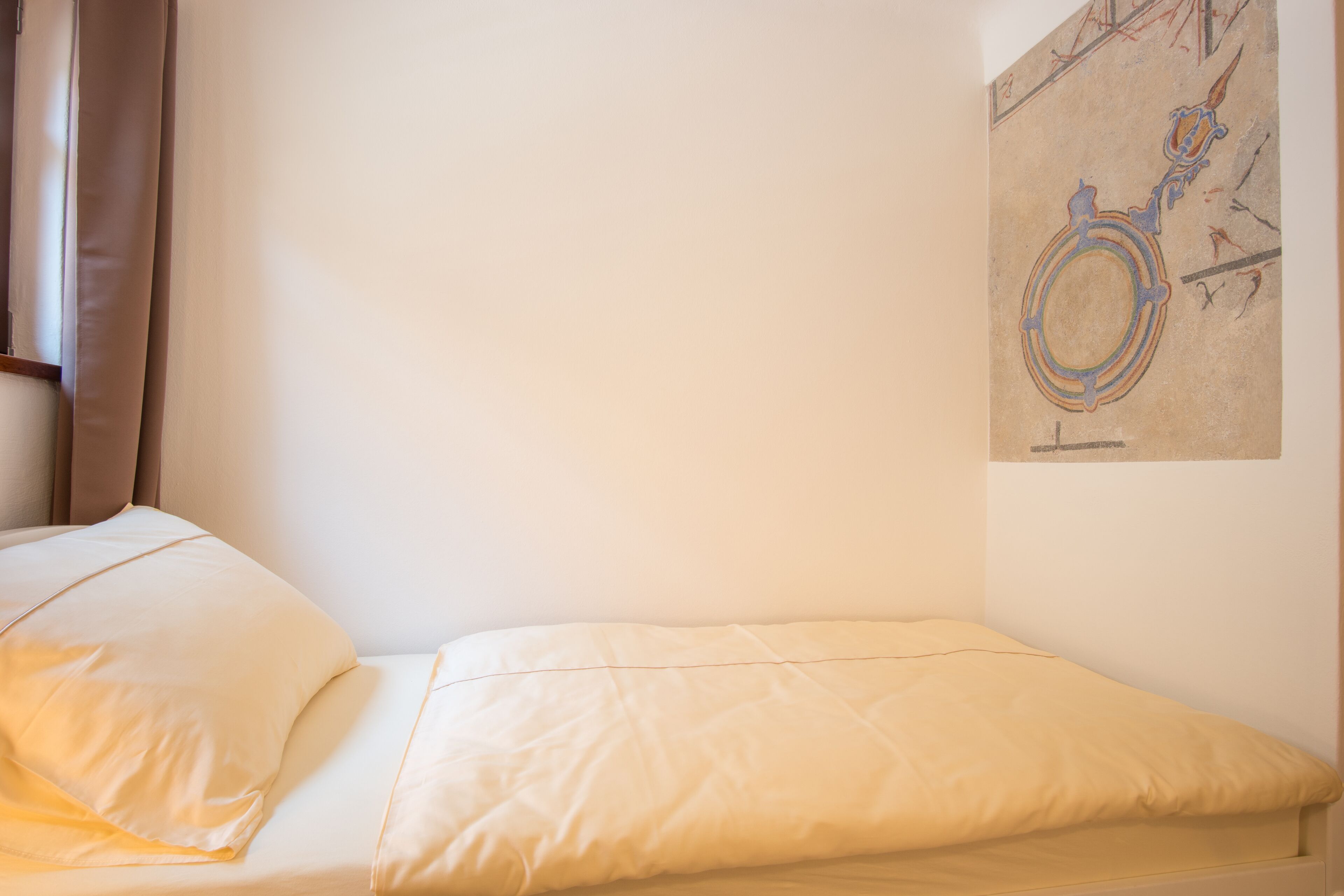Een eenpersoonsbed met een beige matras en kussens, een eenvoudige muur met een kunstwerk met abstracte vormen, en een raam met gordijnen.