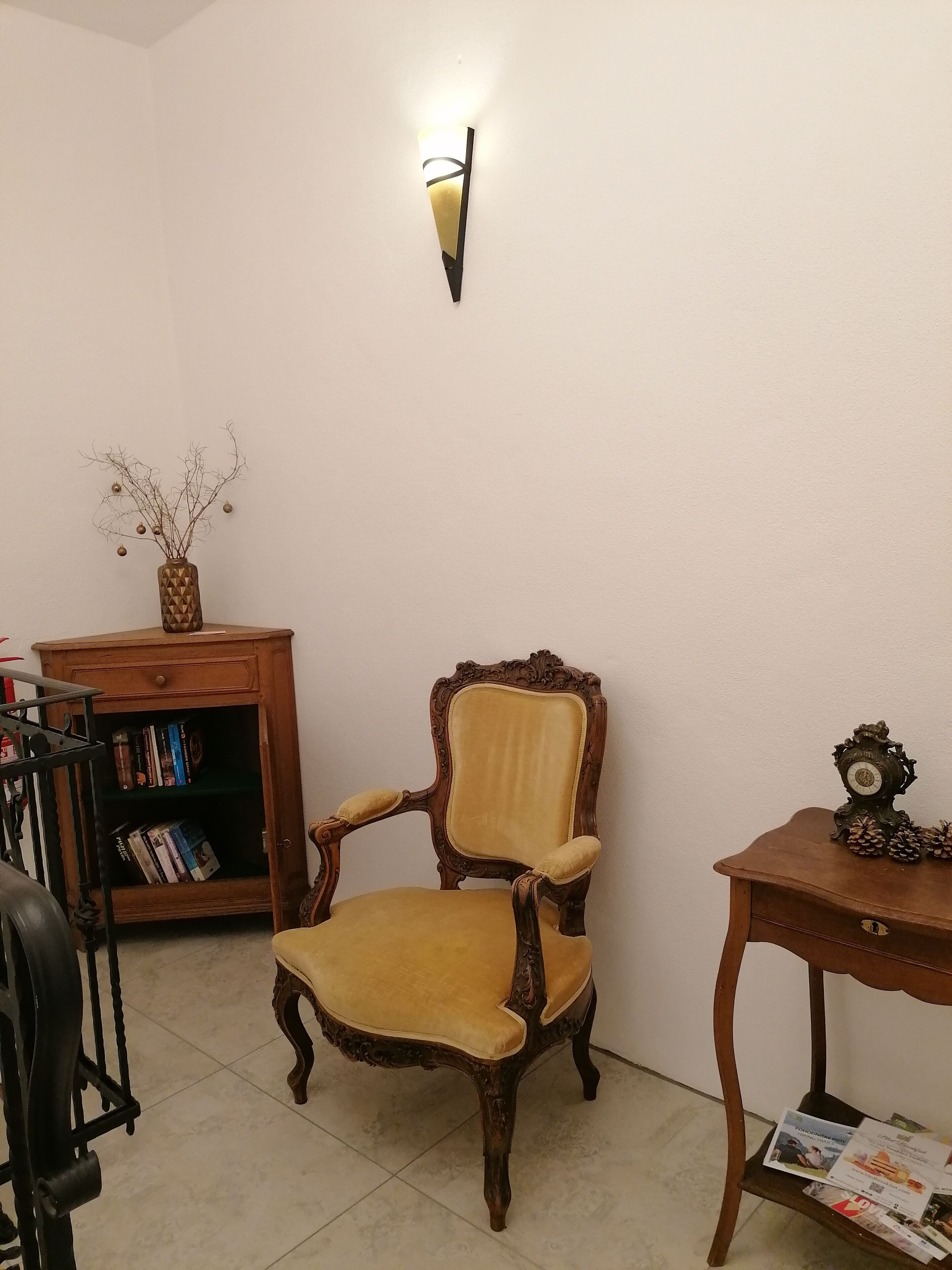 Ein Zimmer mit einem Holzbett, einem kleinen Holztisch mit einer Uhr, einer Wandleuchte und einem Bücherregal mit Büchern.
