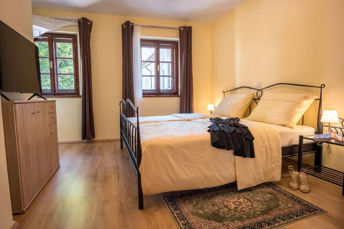 Un dormitorio con cama, mesita de noche, televisión, armario y ventana con cortinas.