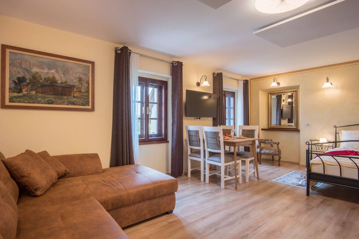 Una sala de estar moderna con un sofá marrón, una mesa de comedor con sillas, un televisor montado en la pared y una cama con una manta roja.