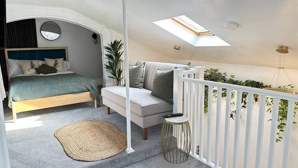 A bedroom with a bed, a small sofa, a round side table, a white railing, and a skylight. - Cosy and Stylish Studio Apartment
 (Trowbridge)