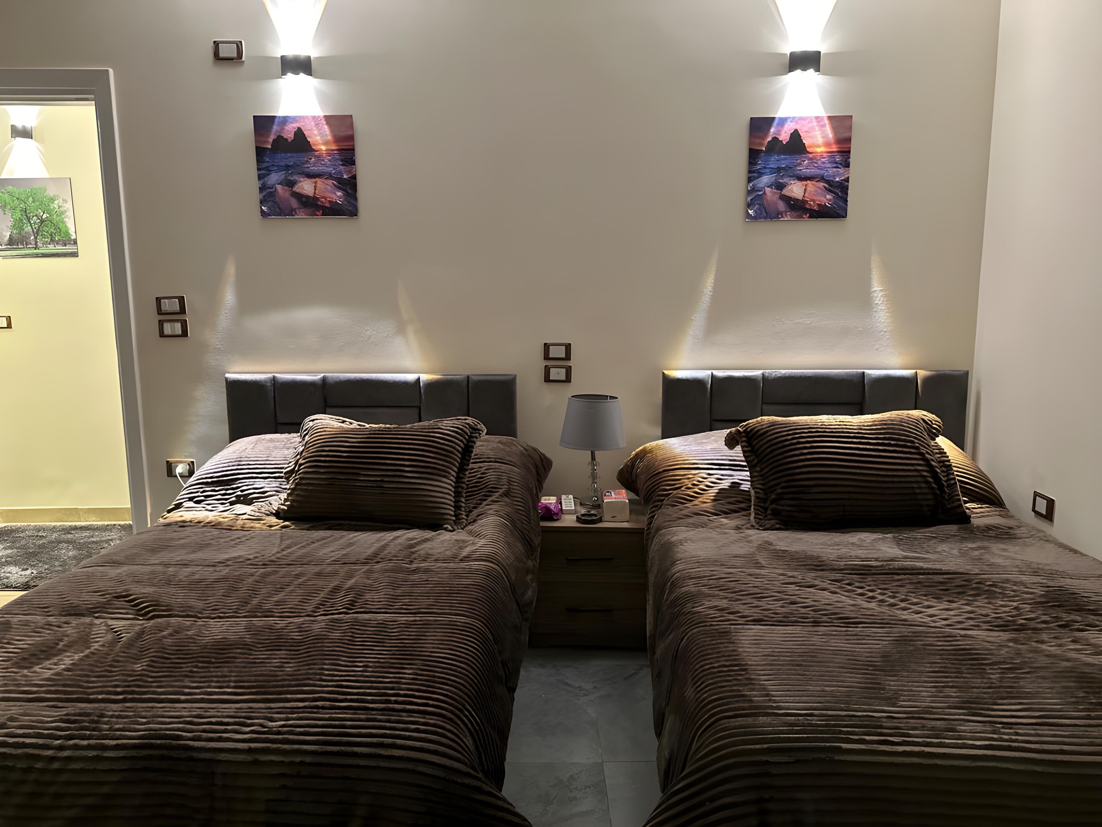A twin bedroom with two beds, each with a headboard, a bedside table with a lamp, and two wall-mounted pictures.