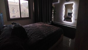 A dimly lit hotel room with a large bed, a window with curtains, and a wall-mounted mirror. - City Center: Modern 2BR Flat-56 (Cairo Governorate)